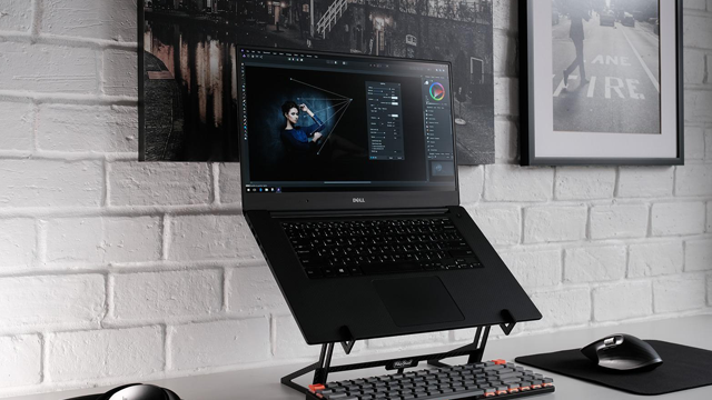 Een zwarte Dell-laptop staat op een bureau met een toetsenbord en muis, tegen een witte bakstenen muur met foto's.