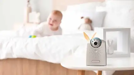 Babyfoon met camera op een witte tafel, baby en knuffel op de achtergrond in een witte kamer.