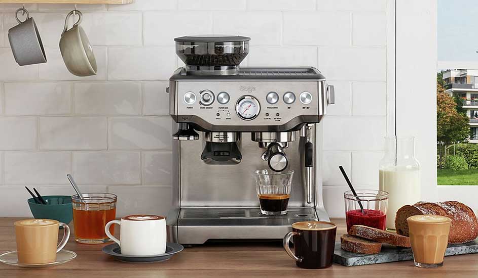 Een zilveren espressomachine staat op het aanrecht, omringd door kopjes koffie, thee, melk, brood en jam in een lichte keuken.