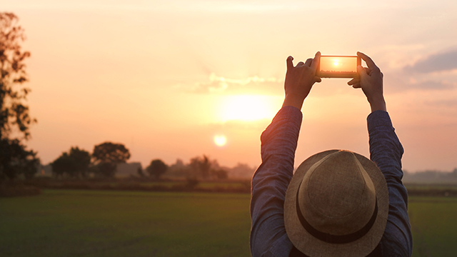 Een persoon met hoed filmt zonsondergang met een telefoon, veld en bomen op achtergrond.