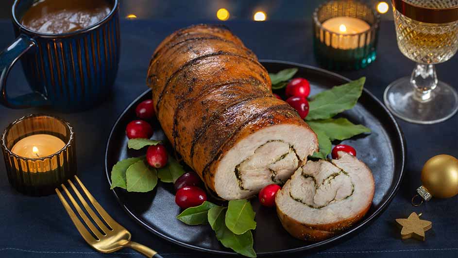 Een gevulde rollade op een zwart bord met rode bessen en groene bladeren, omringd door een beker, kaarsen en een wijnglas.