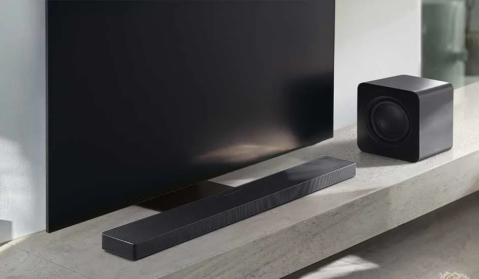 Een zwarte soundbar en subwoofer staan op een lichtgrijze plank direct onder een grote flatscreen tv in een moderne, minimalistische woonkamer.