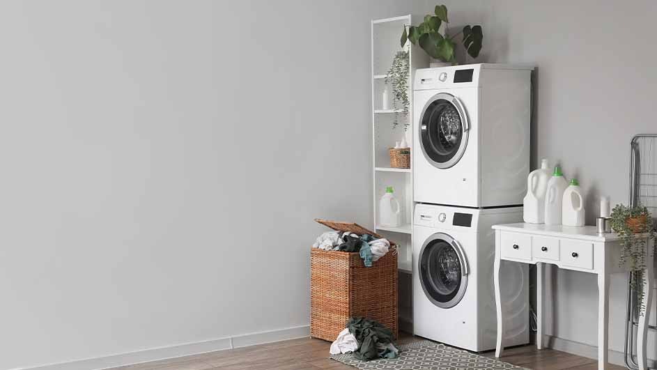 Laundry room with stacked white washer and dryer, wicker basket, white shelf, bottles, and table against a gray wall.