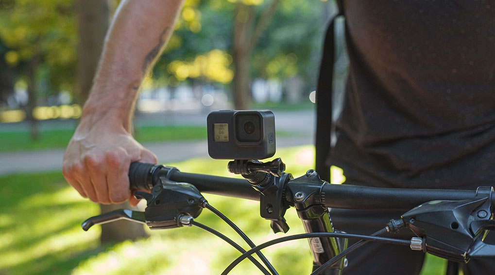 Hand op zwart fietsstuur met GoPro-camera bevestigd, tegen een groene, wazige achtergrond.