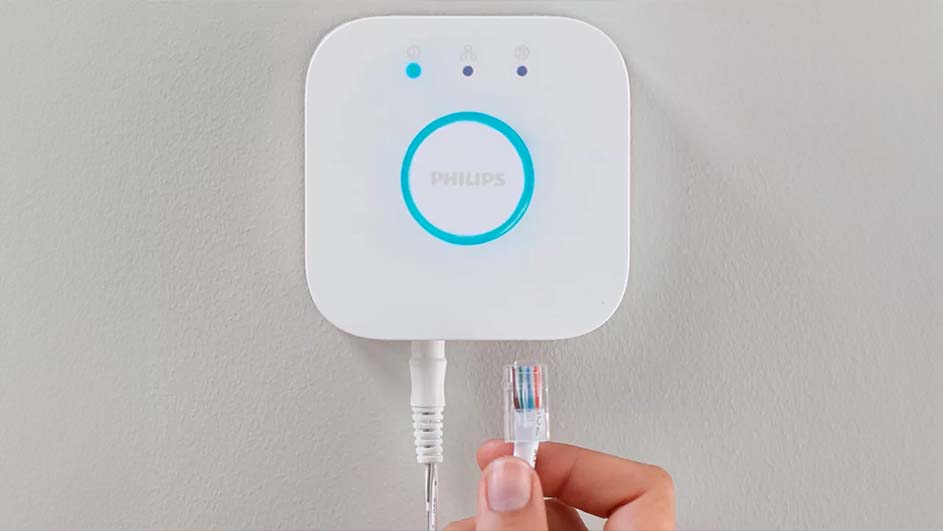 Witte Philips Hue-bridge aan een grijze muur, met blauwe accenten en LED-indicatoren. Een hand sluit een kabel aan.