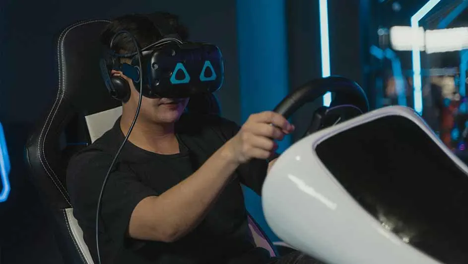Een persoon met een VR-headset zit in een zwarte stoel en houdt een zwart stuur vast in een blauwe omgeving.
