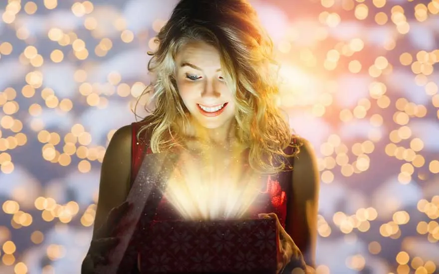 Een verraste jonge vrouw opent een doos waaruit warm magisch licht straalt, omringd door zachte, gouden feestlichtjes op de achtergrond.