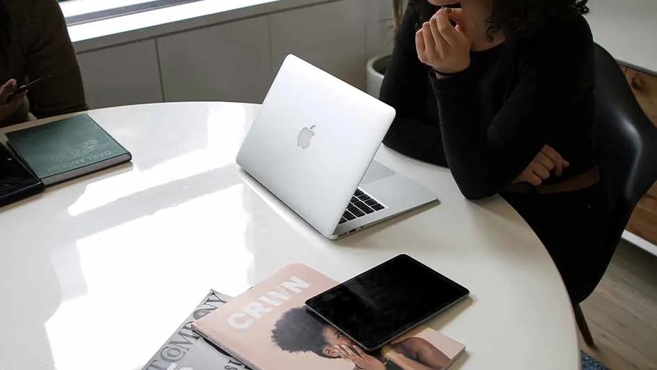 Een witte tafel met een laptop, een tablet, tijdschriften, een notitieboek en twee mensen.