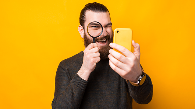Een man met een baard en een grijze trui houdt een vergrootglas voor een oog en een gele telefoon in zijn hand, tegen een gele achtergrond.