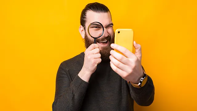 Een man met een baard en een grijze trui houdt een vergrootglas voor een oog en een gele telefoon in zijn hand, tegen een gele achtergrond.