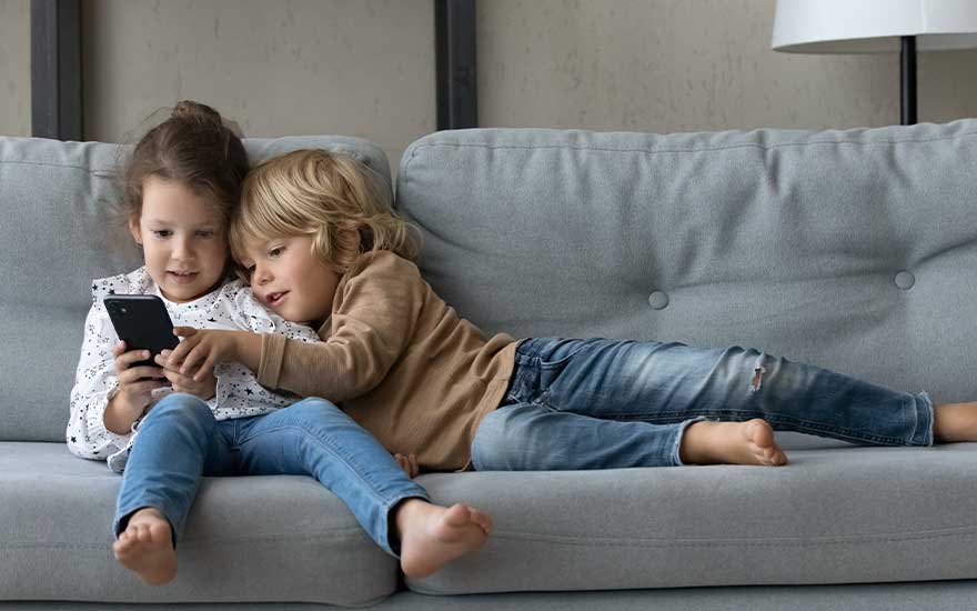Two young children lie comfortably on a grey sofa, closely sharing a smartphone and glimlachend terwijl ze het scherm bekijken.