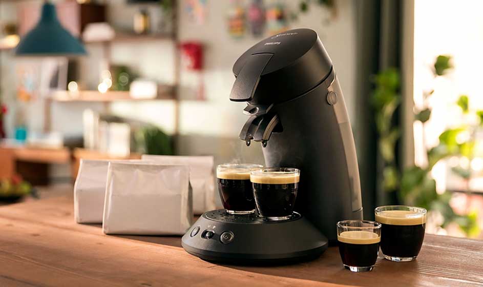 Een zwarte koffiemachine zet twee kopjes koffie op een houten tafel, met extra glazen, witte zakken en een warme huiselijke achtergrond.