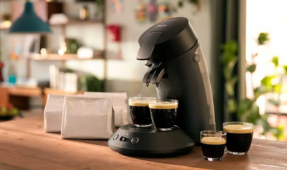 Een zwarte koffiemachine zet twee kopjes koffie op een houten tafel, met extra glazen, witte zakken en een warme huiselijke achtergrond.
