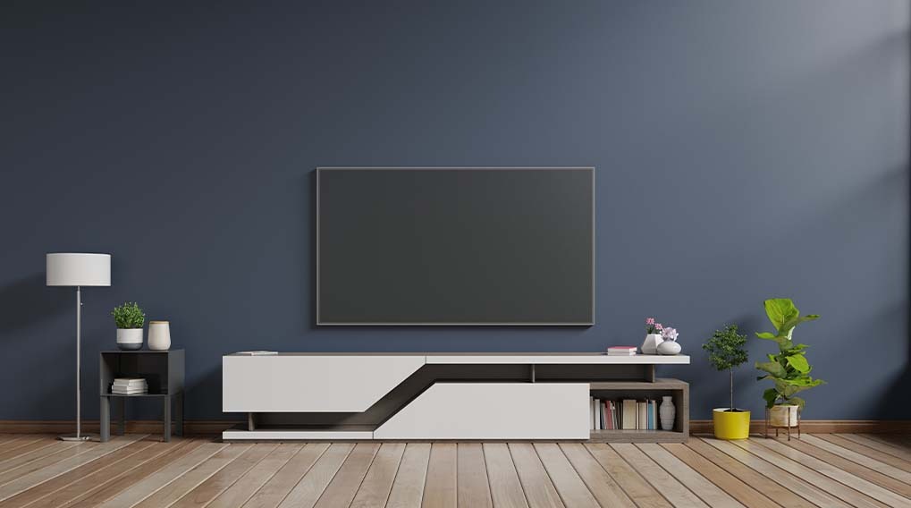 A living room features a large television above a white media console. Plants and a lamp decorate the room. The wall is blue.
