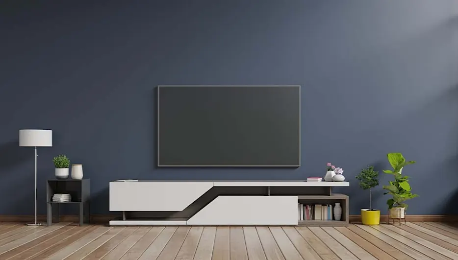 A living room features a large television above a white media console. Plants and a lamp decorate the room. The wall is blue.