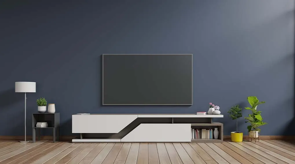 A living room features a large television above a white media console. Plants and a lamp decorate the room. The wall is blue.