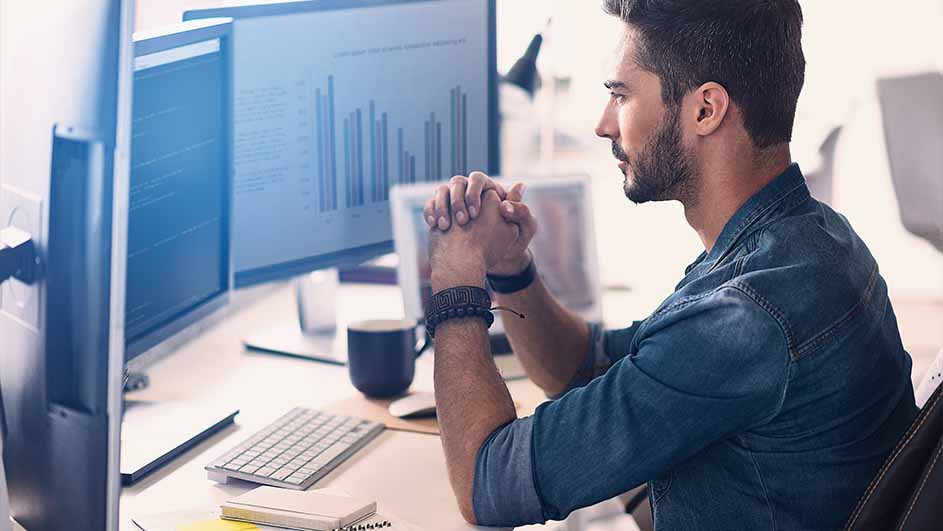 Een man zit aan een bureau met computerschermen met grafieken. Een toetsenbord, notitieboek en beker staan op het bureau.