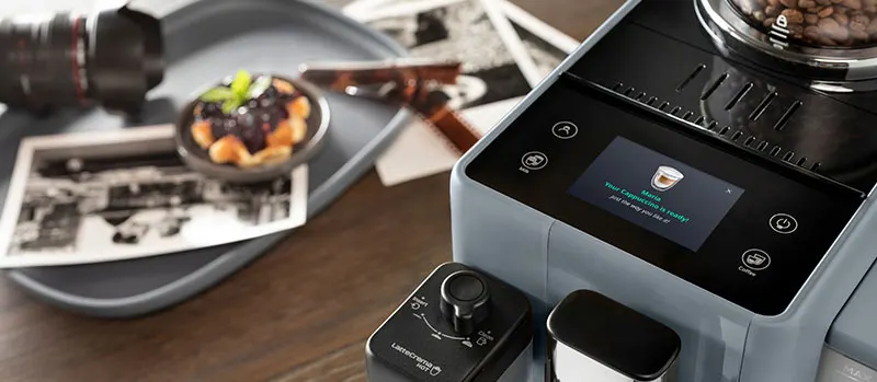 Close-up van een volautomatische espressomachine met touchscreen en koffiebonenreservoir; op de achtergrond liggen foto’s en een bord met dessert.