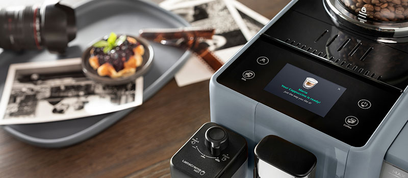 Close-up of a fully automatic espresso machine with a touchscreen and bean hopper, with photos and a dessert plate in the background.