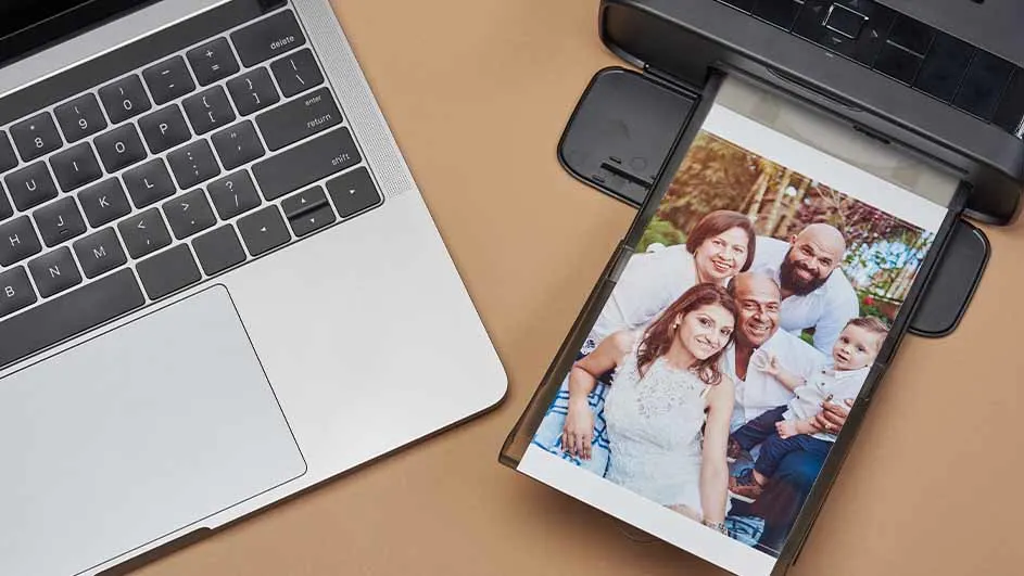Een printer print een foto van een familie, naast een zilveren laptop op een bruine tafel.