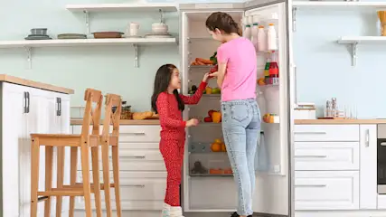 In een keuken staan een vrouw en kind bij een open koelkast vol groenten en flessen.