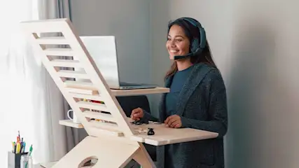 Vrouw met headset werkt aan een houten bureau met laptop. Op het bureau staan pennen en een kop. Gordijnen op de achtergrond.