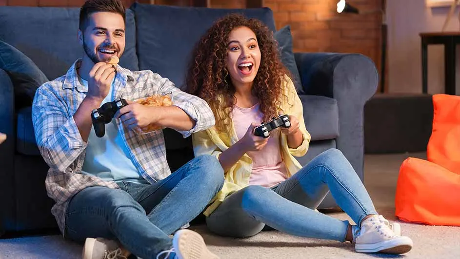 Een man en een vrouw zitten op de vloer, gamen met controllers en eten snacks voor een blauwe bank.
