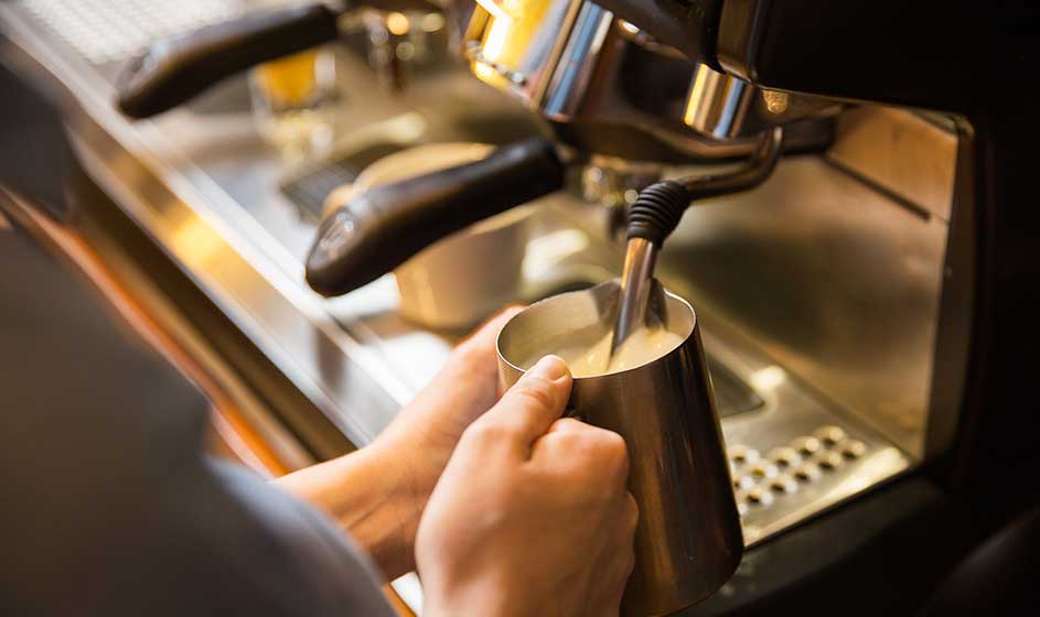 Iemand schuimt melk op in een metalen kannetje bij een espressomachine, met warme verlichting en glanzende onderdelen op de achtergrond.