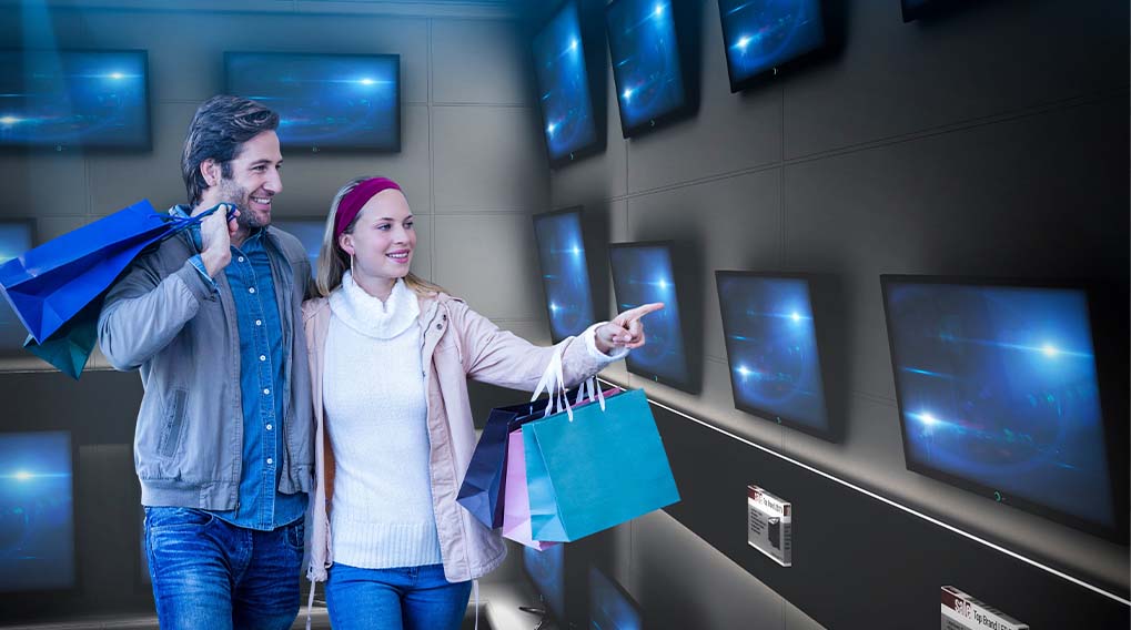 Een man en een vrouw met boodschappentassen kijken naar televisies aan een donkere muur.