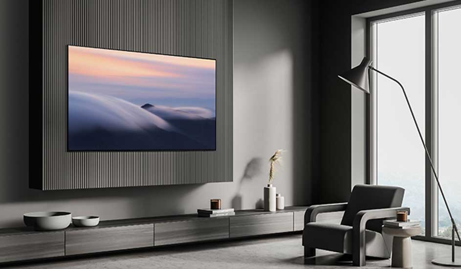 Moderne woonkamer met een grote tv aan de muur, een grijze stoel, lage kast, designlamp en grote ramen met veel licht.