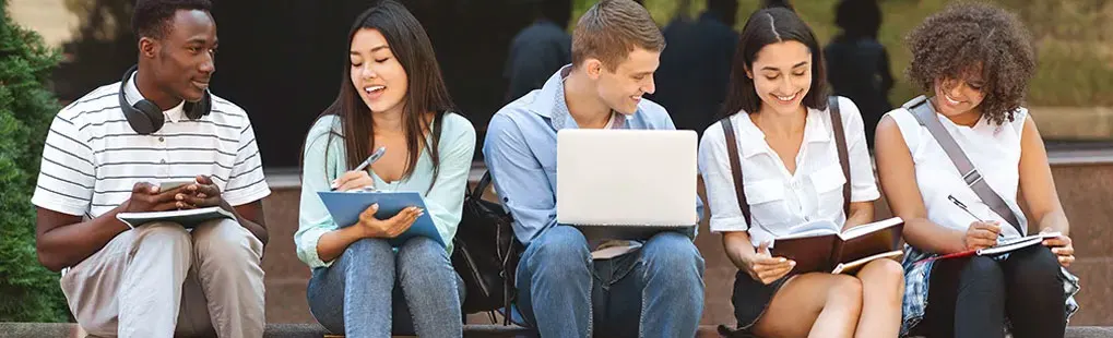 Five students sit outside with notebooks, a laptop, and books. Foliage and people are in the background.