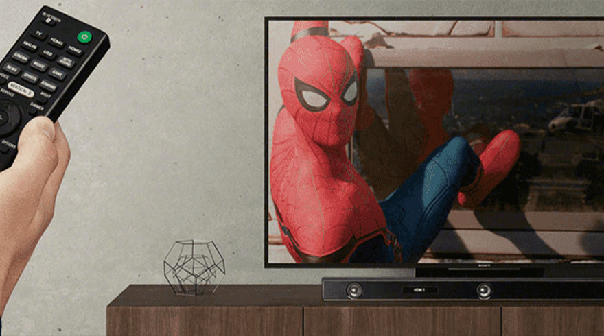 Een hand houdt een zwarte afstandsbediening vast. Een tv toont Spider-Man, staand op een houten kast met een soundbar en een metalen decoratie.