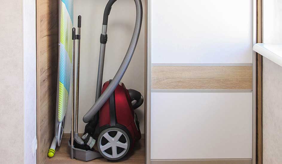 Een rode stofzuiger staat in een smalle kast naast strijkplank en steel, netjes opgeborgen en klaar voor de volgende poetsbeurt.