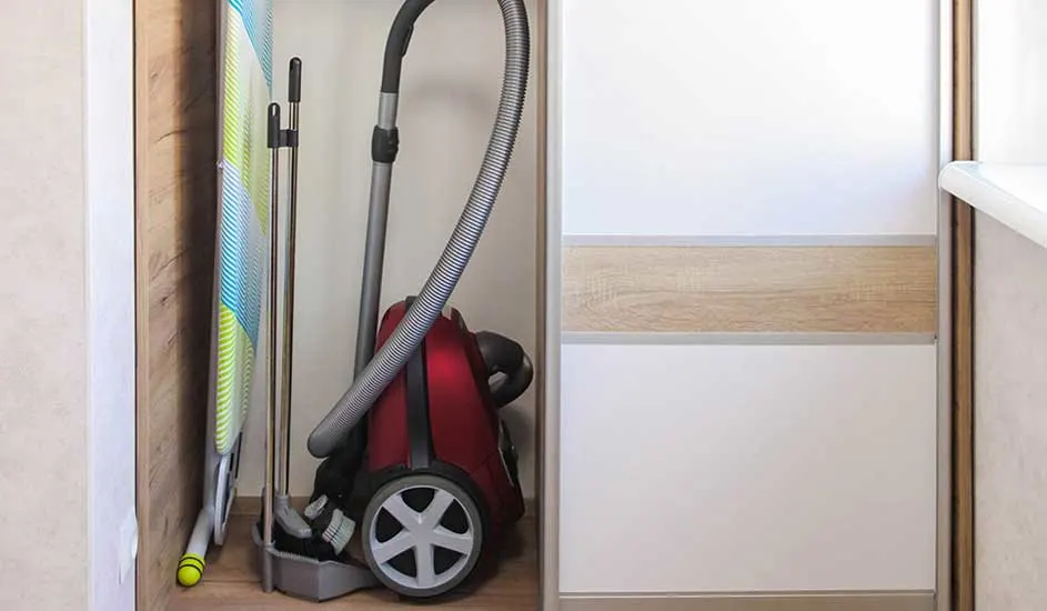 Een rode stofzuiger staat in een smalle kast naast strijkplank en steel, netjes opgeborgen en klaar voor de volgende poetsbeurt.