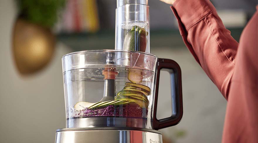 Een keukenmachine met gesneden groenten, waaronder courgette en rode kool, staat klaar voor gebruik. Iemand in een rood shirt staat erbij.