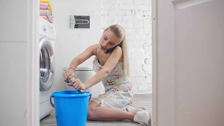 Een vrouw knielt bij een wasmachine met een blauwe emmer, wringt kleding uit en belt met een telefoon in een badkamer.