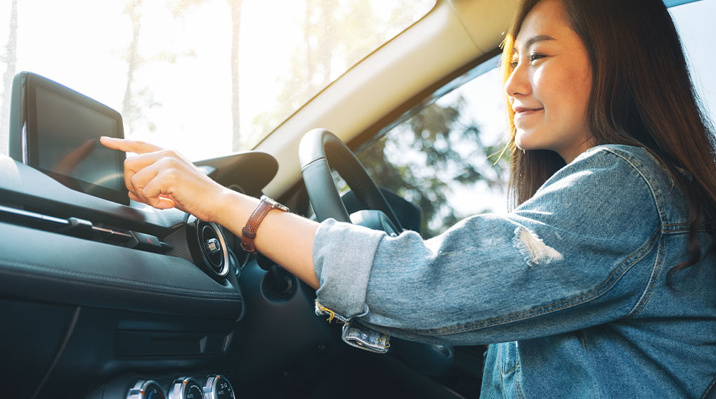 Een vrouw in een auto, die een scherm aanraakt. De auto heeft een zwart dashboard en een stuur.