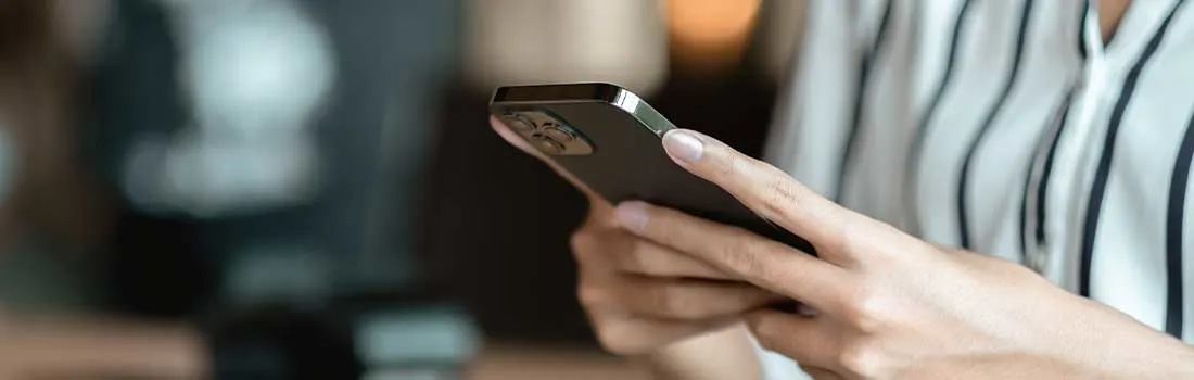 Handen houden een zwarte smartphone vast in een gestreepte blouse, met een onscherpe achtergrond van warme tinten.