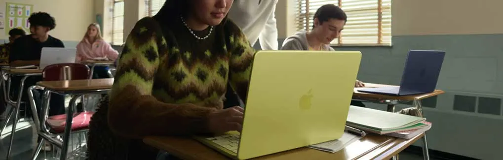 Student works intently on a yellow laptop in a bright classroom, as the teacher leans over to help and classmates focus at nearby desks.