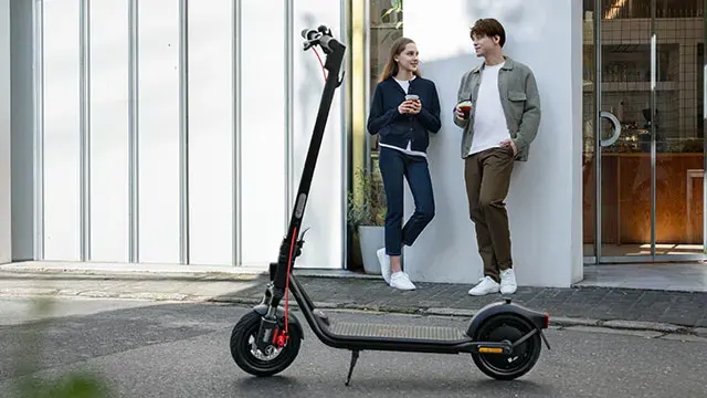 Een zwarte elektrische scooter staat geparkeerd voor een gebouw, met twee mensen die koffie drinken.