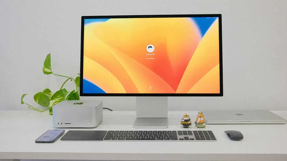 Een bureau met een computer, toetsenbord, muis, telefoon, laptop, plant en decoratieve figuren tegen een witte achtergrond.