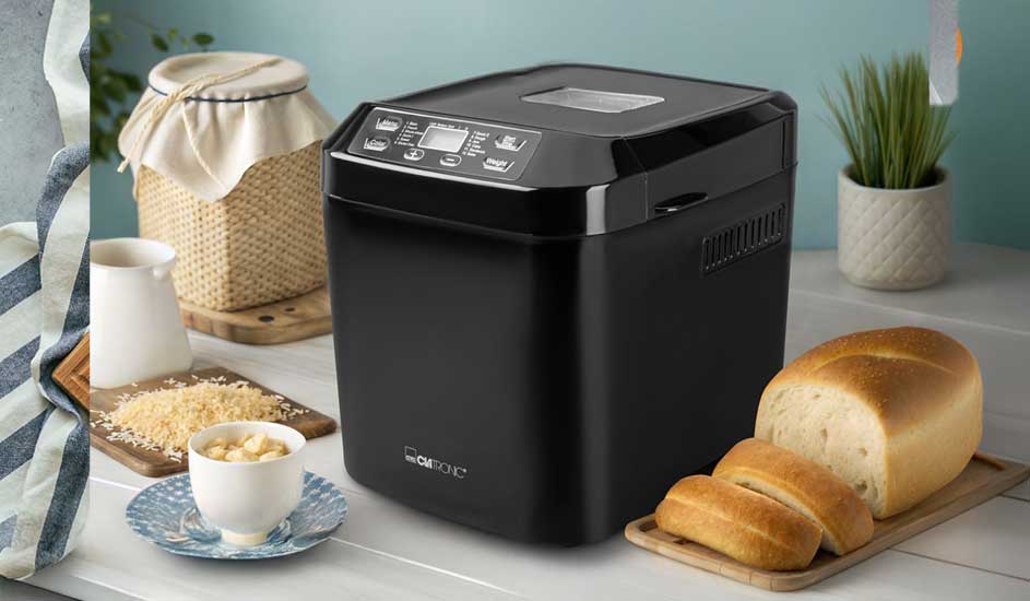 A black bread maker stands on a white table, beside sliced white loaves, small bowls of ingredients and a little green plant.