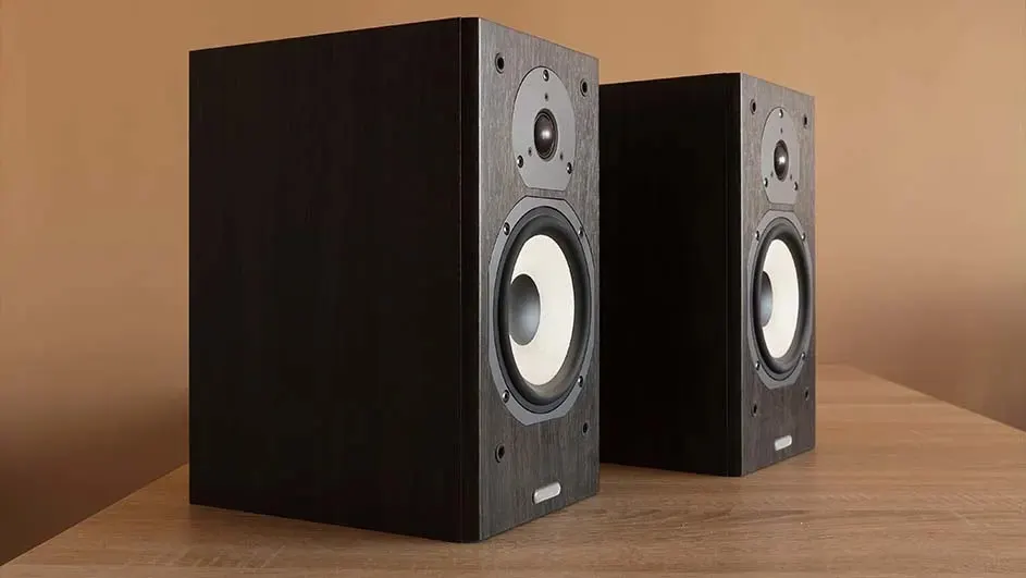 Two rectangular speakers sit on a wooden surface. The speakers are dark, with circular elements on the front. A tan wall is in the background.