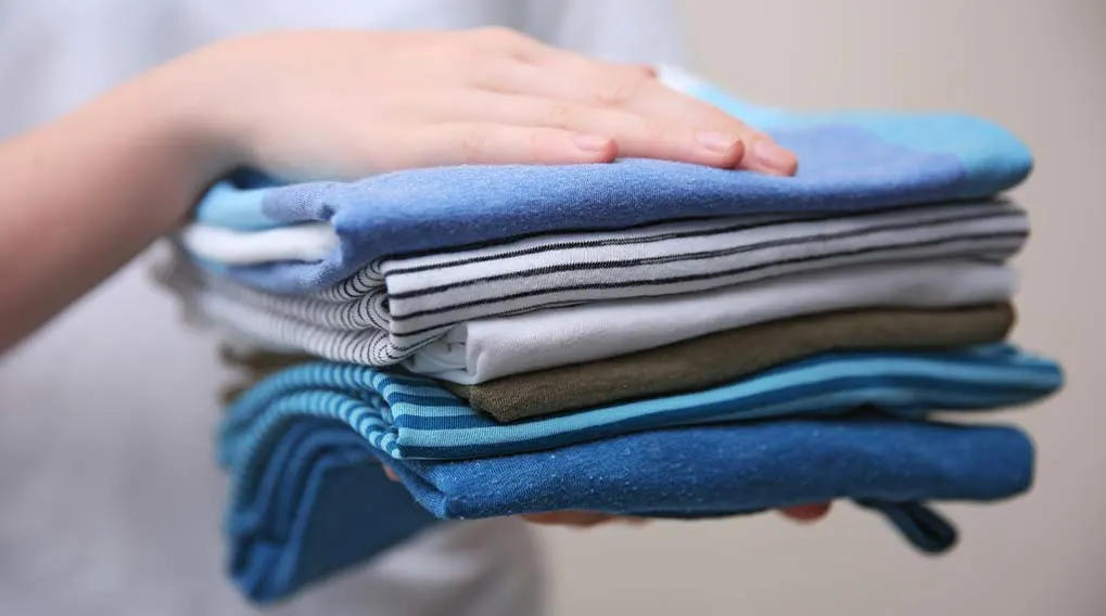 Een hand ligt op een stapel opgevouwen kleding, met blauwe, gestreepte, witte en groene stoffen zichtbaar.