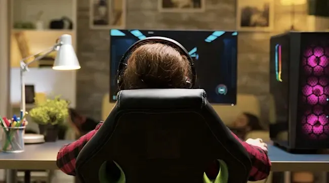 Gamer in ergonomische stoel met headset voor monitor, naast paars verlichte pc-tower, bureau met lamp en plant op achtergrond.