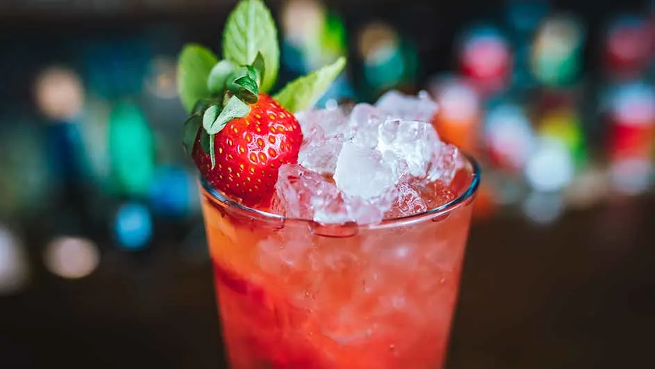 Een glas met een rood drankje gevuld met ijsblokjes, gegarneerd met een aardbei en groene bladeren. Vage lichten op de achtergrond.