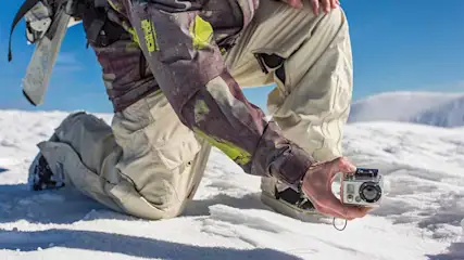 Een persoon knielt in de sneeuw met een ski en een camera in de hand, tegen een achtergrond van een besneeuwde berg.
