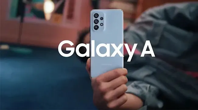 Een hand houdt een lichtblauwe Samsung Galaxy A smartphone omhoog tegen een gezellige woonkamerachtergrond met warme verlichting.