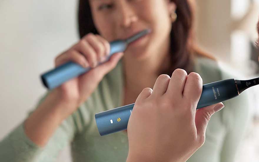 A woman brushes her teeth before a mirror with a blue sonic toothbrush, while her hand is sharply focused in front.