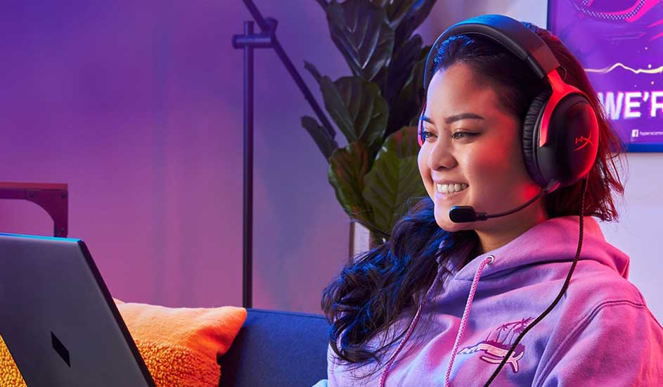A smiling woman in a lilac hoodie wears a gaming headset, chatting at her laptop on a sofa lit by soft pink and blue neon.